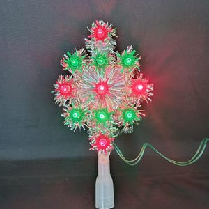 May include: A Christmas tree topper with a starburst design. It has red and green lights, surrounded by silver tinsel. The base is a clear plastic sleeve, and green wires extend from the base.