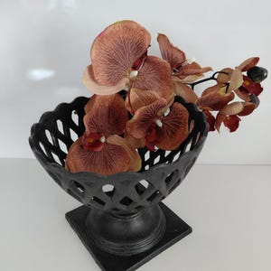 May include: A black decorative bowl with a lattice design, filled with brown and red orchids. The bowl sits on a square base, creating a striking contrast against the white background. The arrangement is a stylish centerpiece.