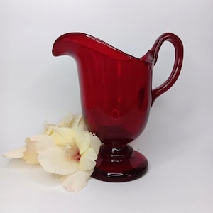 May include: A deep red glass jug with a handle and a flared spout. The jug stands on a pedestal base. Some white and yellow flowers are placed near the base of the jug.