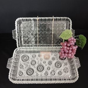May include: Two clear glass serving trays with decorative edges. The top tray has a geometric pattern, while the bottom tray features a circle design. A bunch of purple grapes with green leaves rests on the side, suggesting a serving theme.