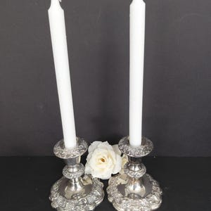 May include: Two silver-toned metal candle holders with intricate embossed floral patterns on the circular bases and rims. Each holder contains a tall, slender, solid white taper candle. A single white artificial rose is positioned between the two holders.
