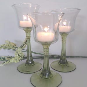 Peut inclure: Trois bougeoirs en verre avec des bougies blanches allumées. Les bougeoirs ont une tige et une base vertes, avec une coupe en verre transparent pour contenir la bougie. Les bougies sont allumées, créant une lueur chaleureuse.
