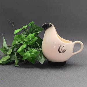 May include: White ceramic creamer with a black rim and a black leaf design. The creamer is shaped like a bird and has a handle.