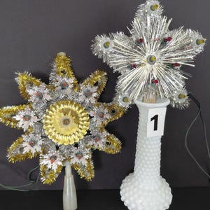 May include: Two star-shaped Christmas tree toppers. One is silver with multi-coloured lights, resting in a white hobnail vase labelled 1. The other is gold and silver with a central gold medallion and lights, labelled 2, featuring a plastic base.