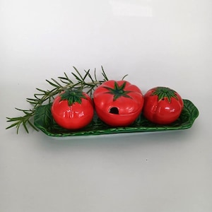 May include: A ceramic tomato-shaped salt and pepper set with a matching lidded container, presented on a green leaf-shaped tray. The tomatoes are bright red with green accents, and rosemary sprigs add a natural touch.