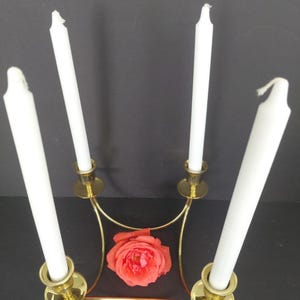 Peut inclure: Un candélabre en métal doré doté de quatre bougeoirs verticaux disposés en carré. Chaque support contient une longue bougie effilée blanche. Une rose unique de couleur corail vif est placée au centre de la structure métallique.