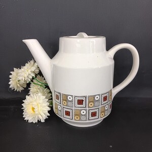 May include: White ceramic teapot with a lid and handle. The teapot has a geometric pattern of squares and circles in brown, gold, and black. A white spout extends from the top. Artificial white flowers are on the left side of the teapot.