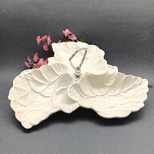 May include: White ceramic serving dish in a leaf design with a silver handle. The dish has three sections and is decorated with leaf-like textures. Pink flowers are in the background.