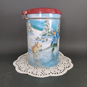 May include: A red and white tin container with a winter scene featuring a boy playing hockey with a dog. The tin has a red lid with a metal handle.
