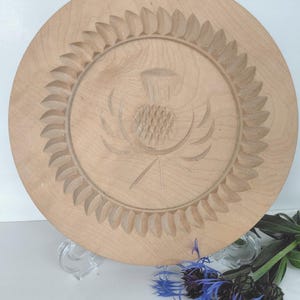 May include: A round, light-coloured wooden plate featuring a carved thistle design at its centre. The plate has a decorative border of carved leaf shapes. Blue flowers are displayed with the plate.