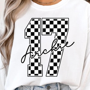 May include: White long-sleeve sweatshirt featuring the number 17 in a black and white checkered pattern. The name "Archie" is written in a cursive font below the number. The sweatshirt is a casual style.