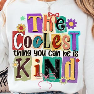 May include: White sweatshirt with the text "The Coolest thing you can be is Kind" in colorful, cut-out style letters. The design includes flowers and bows. A person is wearing the sweatshirt and giving a peace sign.