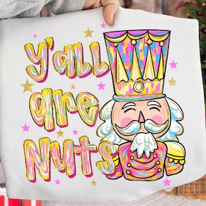May include: White sweatshirt featuring the phrase "Y'all are Nuts" in colourful bubble letters. A cartoon nutcracker with a crown and uniform is depicted. Gold and pink stars are scattered around the text and image.