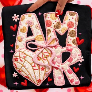 May include: Black t-shirt with the word "AMOR" embroidered in a decorative font. The "A" and "M" are filled with pastry-like designs, while the "R" is filled with hearts. A pink bow and heart-shaped design are also present.