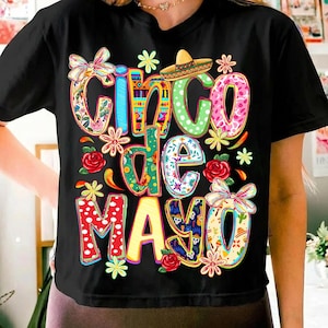 May include: Black t-shirt with a colorful "Cinco de Mayo" design. The design includes floral patterns, a sombrero, and the text in various colors. The shirt is adorned with flowers and bows.