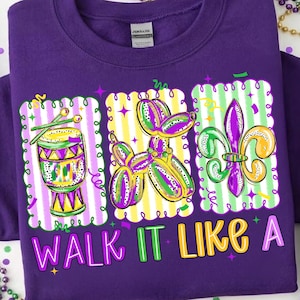 Puede incluir: Sudadera morada con un diseño gráfico colorido que presenta un tambor, un animal de globo y una flor de lis. El texto "WALK IT LIKE A" está impreso en la parte inferior en morado, amarillo y verde. El diseño tiene un tema festivo.