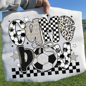 May include: White soccer-themed blanket with the words "Game Day" in a mix of fonts and patterns. The letters feature soccer ball designs, checkered patterns, and leopard print. A soccer ball with a bow is in the center.