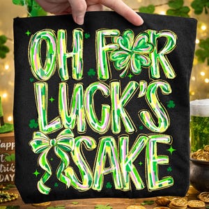 Puede incluir: Bolso tote negro con el texto "OH FOR LUCK'S SAKE" en letras verdes y amarillas, con detalles de tréboles y lazos. El bolso es sostenido por una mano, con temática del Día de San Patricio.