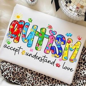May include: White t-shirt with the word "AUTISM" in colorful puzzle piece letters, decorated with flowers and handprints. The words "accept, understand, love" are below the main text.