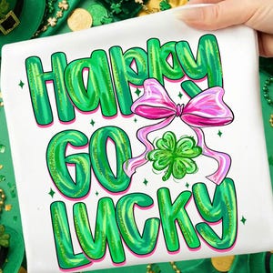 Könnte beinhalten: Weißes T-Shirt mit den Worten "HAPPY GO LUCKY" in grünen Glitzerbuchstaben. Eine rosa Schleife und ein grünes Kleeblatt sind im Design enthalten. Das T-Shirt wird von einer Hand gehalten, mit grünen St. Patrick's Day Dekorationen im Hintergrund.