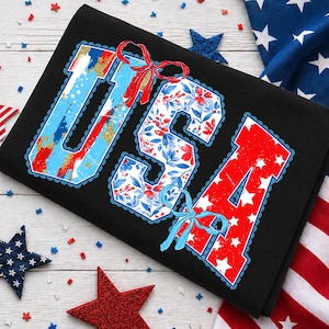 May include: Black t-shirt with the word "USA" in red, white, and blue. The letters are decorated with floral and patriotic patterns, and the design includes bows. The shirt is surrounded by star-shaped decorations and an American flag.