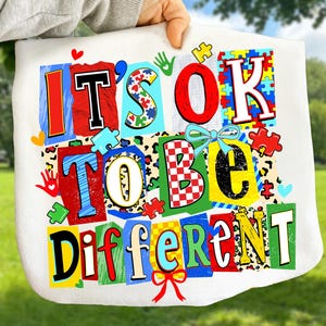 May include: White tote bag with the text "IT'S OK TO BE DIFFERENT" in colorful, block letter style. The design includes puzzle pieces, handprints, and bows. The background is a blurred green.