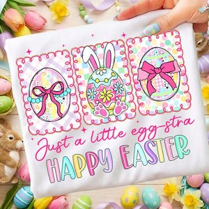 May include: White t-shirt with three Easter egg designs and the text "Just a little egg-stra HAPPY EASTER" in colorful letters. The eggs are decorated with bows, bunny ears, and floral patterns, all framed in pink.