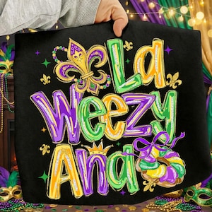 May include: Black tote bag with the text "La Weez Ana" in yellow, green, and purple. The design includes a fleur-de-lis, a king cake, and stars. The bag is held by a person.