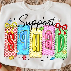 May include: White sweatshirt with the words "Support Squad" in black script. The word "Squad" is spelled out in colorful, patterned blocks. Each letter is a different color and design, with puzzle pieces and floral accents.
