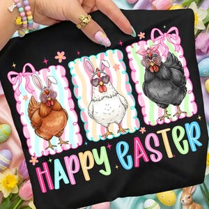 May include: Black t-shirt featuring three cartoon chickens wearing sunglasses and bunny ears, set against a striped background. The words "HAPPY EASTER" are displayed in colorful letters. The design is held by a person.