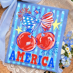 May include: A light blue t-shirt featuring a patriotic design. The shirt displays a pair of red cherries with a bow in the colors of the American flag, surrounded by stars and the word "AMERICA" in bold letters.