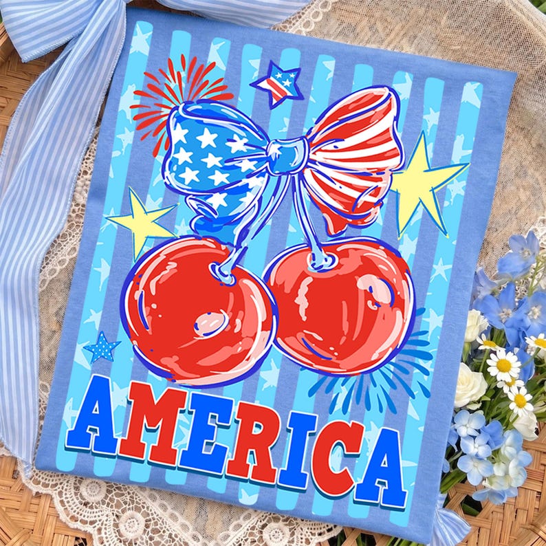 May include: A light blue t-shirt featuring a patriotic design. The shirt displays a pair of red cherries with a bow in the colors of the American flag, surrounded by stars and the word "AMERICA" in bold letters.