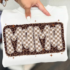 May include: White sweatshirt featuring the word "MAMA" in a retro design. The letters are in a checkered pattern with a brown and tan color scheme, outlined with a string of pearls, and set against a leopard print background. Brown starbursts are scattered around the design.