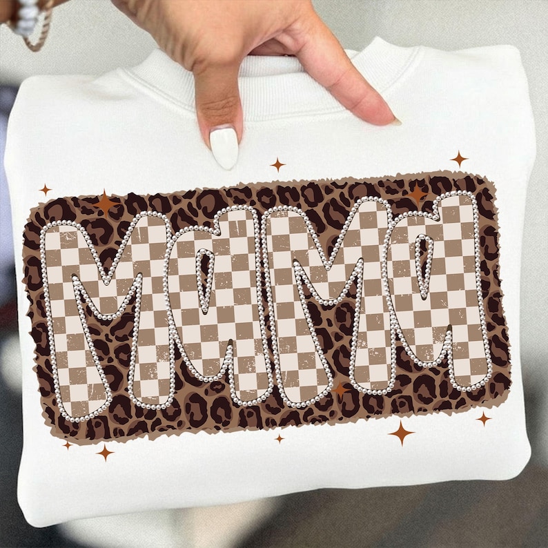 May include: White sweatshirt featuring the word "MAMA" in a retro design. The letters are in a checkered pattern with a brown and tan color scheme, outlined with a string of pearls, and set against a leopard print background. Brown starbursts are scattered around the design.