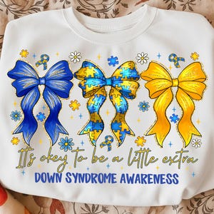 May include: White sweatshirt with three large bows in blue, yellow, and puzzle piece designs. The text "It's okay to be a little extra" and "Down Syndrome Awareness" are printed below the bows. The design includes flowers and puzzle pieces.