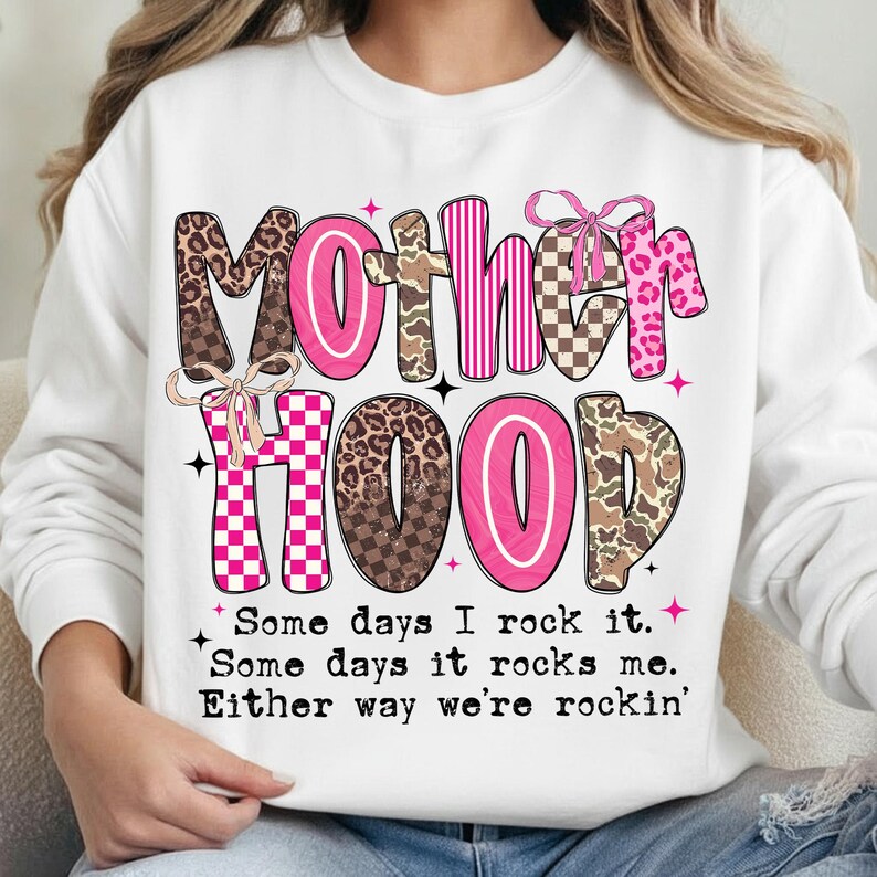 Puede incluir: Sudadera blanca con la palabra "MOTHERHOOD" en una mezcla de letras rosas, marrones y estampadas de animales. El texto "Some days I rock it. Some days it rocks me. Either way we're rockin'" est&aacute; debajo.