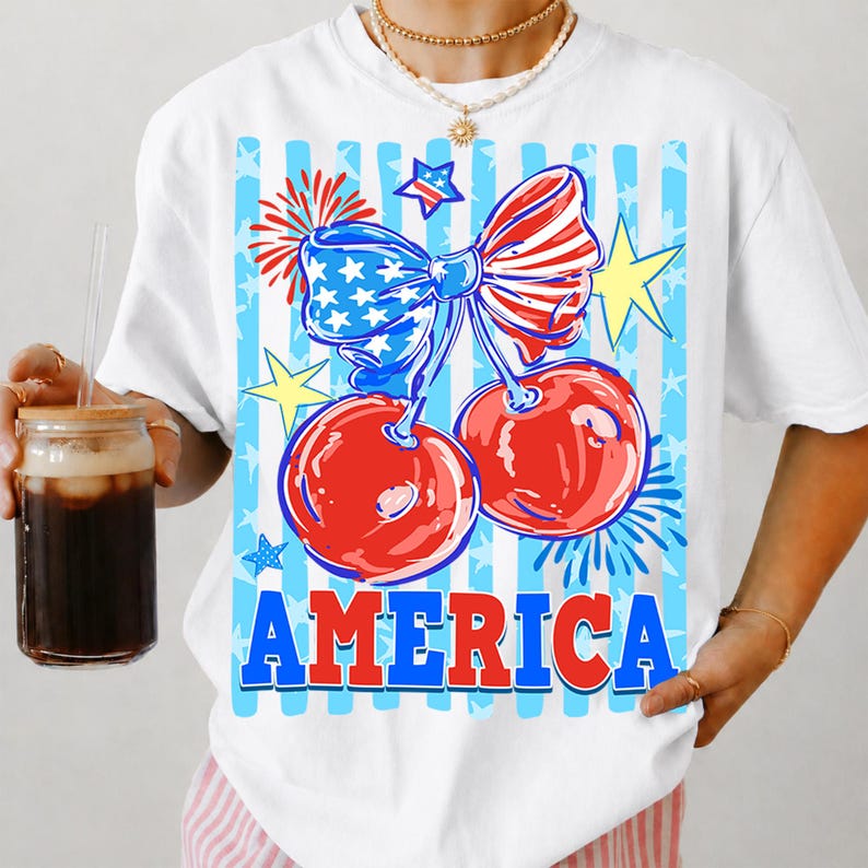 May include: White t-shirt featuring a patriotic design with red cherries, a blue bow with stars, and the word "AMERICA" in bold letters. The design includes blue and white stripes, yellow stars, and fireworks.