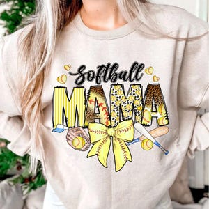 May include: A beige sweatshirt with the text "Softball Mama" in yellow, black, and brown. The text is decorated with a yellow and brown bow, baseballs, and bats.