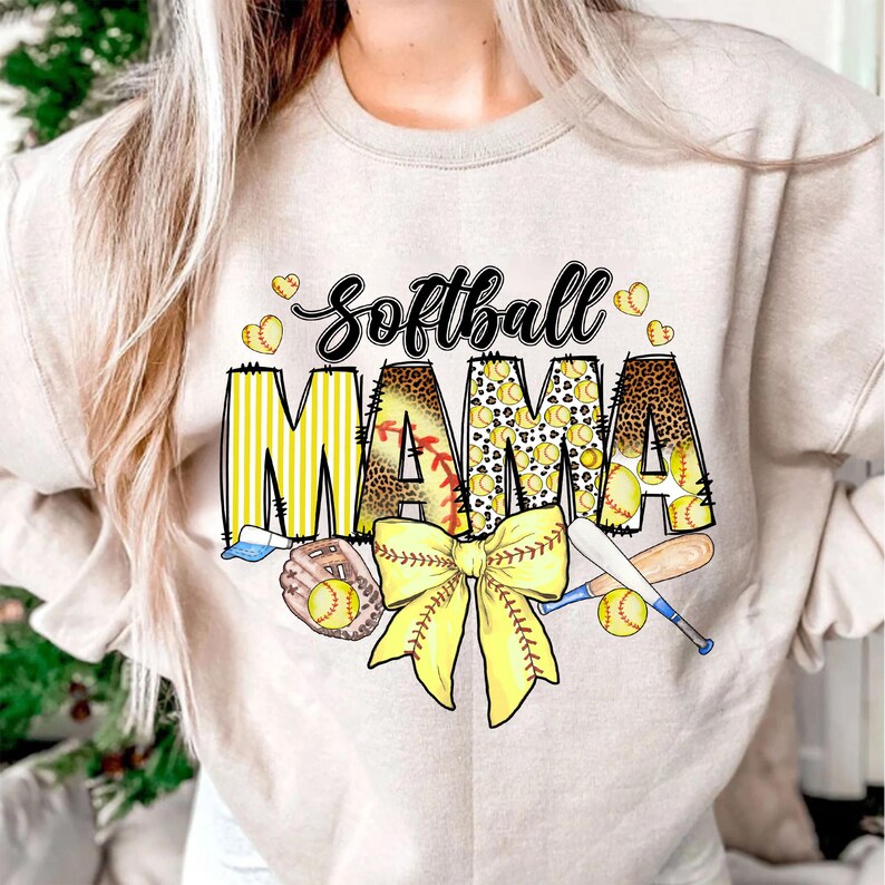 May include: A beige sweatshirt with the text "Softball Mama" in yellow, black, and brown. The text is decorated with a yellow and brown bow, baseballs, and bats.