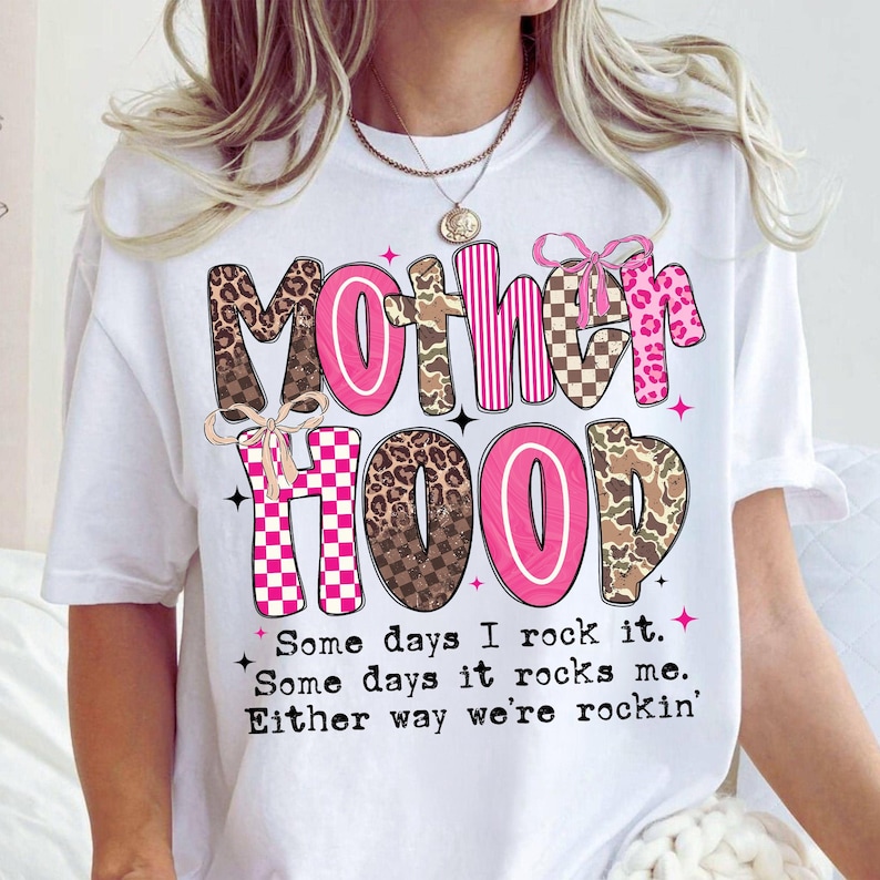 Puede incluir: Camiseta blanca con la palabra "Motherhood" en rosa, marr&oacute;n y fuentes estampadas. La camiseta incluye el texto "Some days I rock it. Some days it rocks me. Either way we're rockin'" en una fuente de escritura.