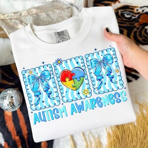 May include: White sweatshirt with a colorful graphic promoting Autism Awareness. The design includes a heart-shaped puzzle, blue bows, and the words "AUTISM AWARENESS" in blue. The sweatshirt is held by a person.