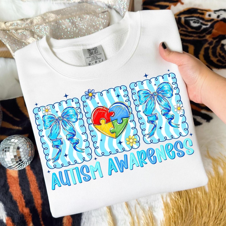 May include: White sweatshirt with a colorful graphic promoting Autism Awareness. The design includes a heart-shaped puzzle, blue bows, and the words "AUTISM AWARENESS" in blue. The sweatshirt is held by a person.