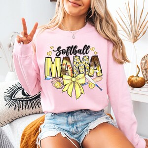 May include: Pink sweatshirt with the text "Softball Mama" in yellow and black letters. The design includes a yellow bow, baseball bats, and baseballs.