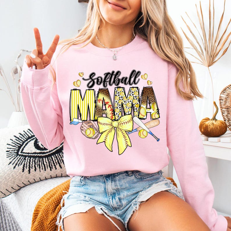 May include: Pink sweatshirt with the text "Softball Mama" in yellow and black letters. The design includes a yellow bow, baseball bats, and baseballs.