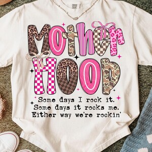 Puede incluir: Camiseta color crema con la palabra "MOTHERHOOD" en una mezcla de rosa, estampado de leopardo, camuflaje y patrones de cuadros. La camiseta tambi&eacute;n presenta el texto "Some days I rock it. Some days it rocks me. Either way we're rockin'".