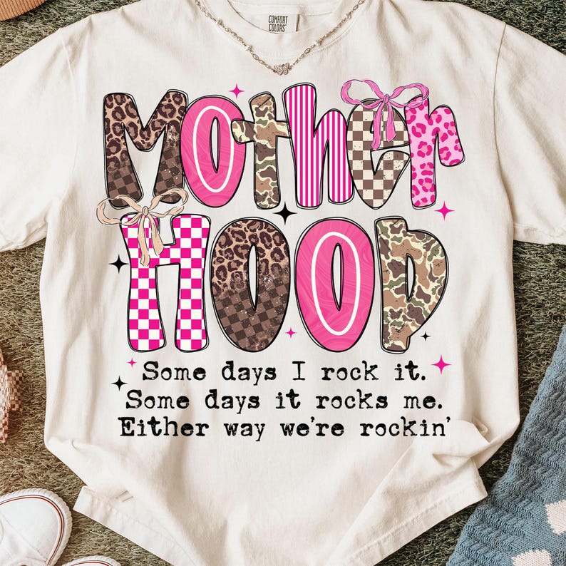 Puede incluir: Camiseta color crema con la palabra "MOTHERHOOD" en una mezcla de rosa, estampado de leopardo, camuflaje y patrones de cuadros. La camiseta tambi&eacute;n presenta el texto "Some days I rock it. Some days it rocks me. Either way we're rockin'".