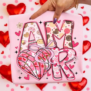 Op de afbeelding: Een lichtroze t-shirt met het woord "AMOR" in een decoratief lettertype. De "A" is gevuld met gebakillustraties, de "M" met hartvormige patronen en de "R" met harten. Een hartvormig ontwerp met een strik staat in het midden.