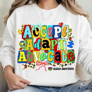 May include: White sweatshirt with the words "Accept, Adapt, Advocate" in colorful, block letters. The design includes puzzle pieces, ribbons, and the text "Autism Awareness".