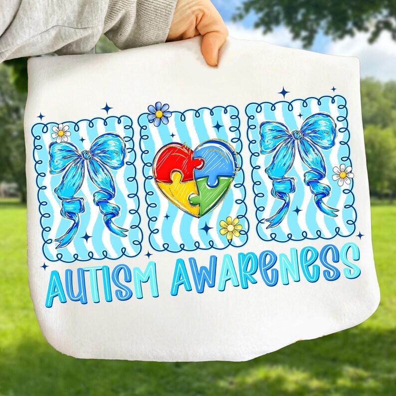 May include: White tote bag with the words "AUTISM AWARENESS" in blue and teal. The design includes blue bows, daisies, and a heart-shaped puzzle piece design in red, yellow, green, and blue. The bag is held up by a person.