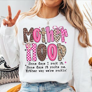 Puede incluir: Sudadera blanca con la palabra "Motherhood" en rosa y letras estampadas. El texto "Some days I rock it. Some days it rocks me. Either way we're rockin'" est&aacute; impreso debajo.
